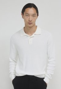 Junger Mann mit langen dunklen Haaren, der ein weißes, gestricktes Langarmpolohemd und schwarze Hosen trägt, mit Händen in den Taschen, neutraler Hintergrund.