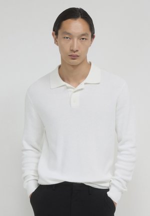 Junger Mann mit langen dunklen Haaren, der ein weißes, gestricktes Langarmpolohemd und schwarze Hosen trägt, mit Händen in den Taschen, neutraler Hintergrund.