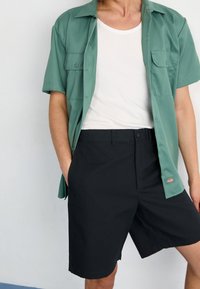 Person som bär svarta shorts, vit linne-tröja och en öppen grön kortärmad skjorta med knappar, med händerna i fickorna.