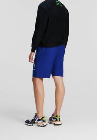 Sudadera negra con logo verde, pantalones cortos azules con logo lateral y zapatillas negras con detalles multicolores y suela gruesa.