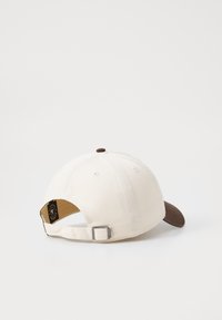 Witte katoenen pet met een bruine klep. Voorzien van een stoffen riem en een metalen gesp aan de achterkant. Ronde top met een kleine bruine knop.