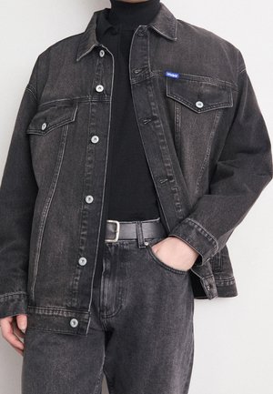 Personne portant une veste en jean noire avec des boutons, un col roulé noir, un jean noir assorti et une ceinture noire avec une boucle argentée.