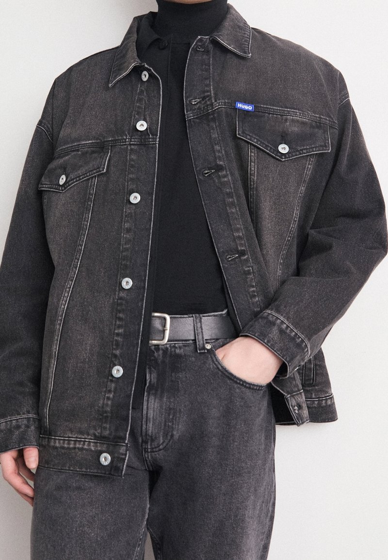 Personne portant une veste en jean noire avec des boutons, un col roulé noir, un jean noir assorti et une ceinture noire avec une boucle argentée.