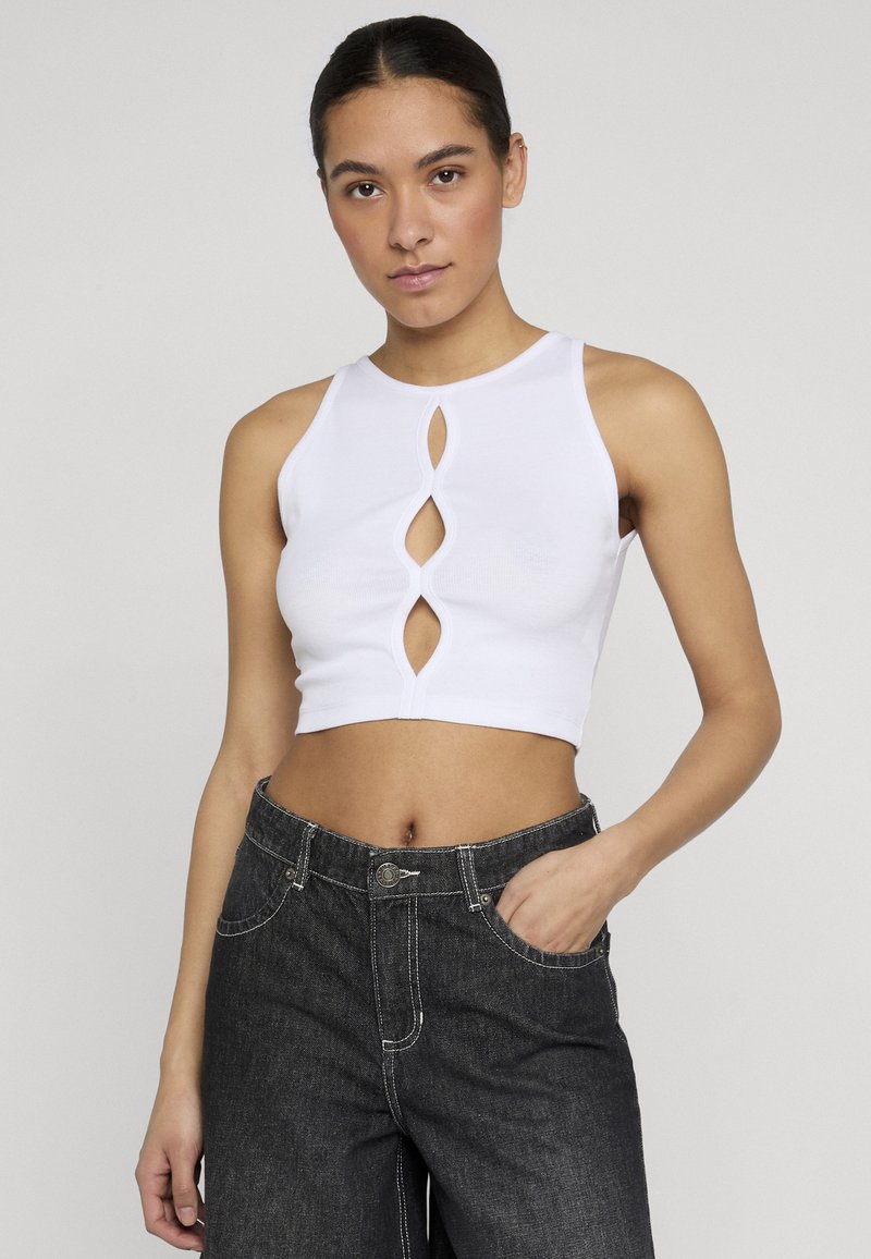 Γυναίκα που φοράει λευκό αμάνικο crop top με τρεις κάθετες εγκοπές και σκουρόχρωμα τζιν με ψηλή μέση, το χέρι ξεκουράζεται στην τσέπη.