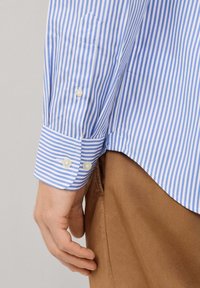 Camisa de manga larga a rayas en azul claro y blanco con puños redondeados, que presenta dos botones, un ajuste entallado y un detalle de bolsillo puntiagudo.