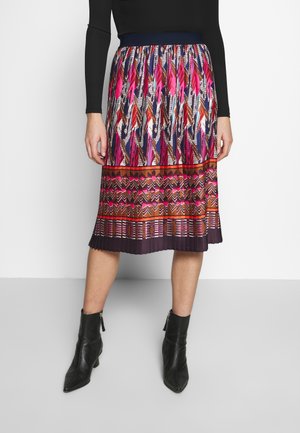 Spódnica plisowana z kolorowymi wzorami piór i geometrycznymi, z ciemnym pasem i fioletowym obszyciem. Noszona z czarnymi botkami do kostki.