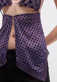 Hõbedaselt lilla täpiline läbipaistev kangas, millel on pitsist ääris allosas ja ees väike kinnitusdetail. Allpool on nähtav must vöö.