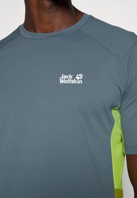 Šedé sportovní tričko s zelenými bočními akcenty, vyrobené z texturované tkaniny. Má kulatý výstřih a bílou logo na hrudi.