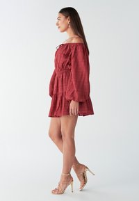 Off-shoulder mini-jurk in roestkleurige tint met een gestructureerde stof, elastische taille en gelaagde ruches. Gecombineerd met gouden sandaalhakken met bandjes.