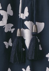 Marinfärgad tyg med vita fjärils mönster. Två mörk marinblå kvastar med metalliska detaljer hänger från en dragsko vid nacklinjen.