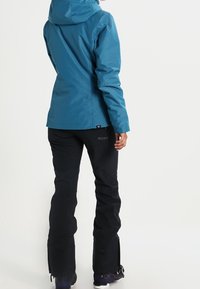 Person in blauer Kapuzenjacke und schwarzen Schneehosen, steht mit dem Rücken zur Kamera vor einem einfarbigen Hintergrund.