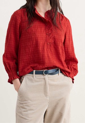 Nainen, jolla on punainen röyhelöinen pitkähihainen pusero beigeihin samettihousuihin tunnettuna mustan vyön kanssa, kädet taskuissa.