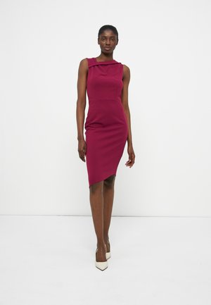 Γυναίκα που φοράει μαν sleeveless, εφαρμοστή, μαγνταλένια φόρεμα μήκους μέχρι το γόνατο με ασύμμετρο τελείωμα και λευκά παπούτσια με τακούνι, στέκεται μπροστά σε απλό λευκό φόντο.