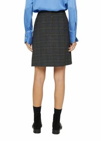 Femme portant un blouse longue à manches bleues, une jupe noire à carreaux arrivant au genou, des bottes noires à la cheville et des chaussettes noires, vue de dos.