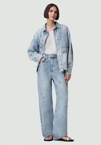 Ljusblå denimjacka och vida jeans med blommönsterbroderi. Vit t-shirt under. Platta sandaler fulländar outfiten.