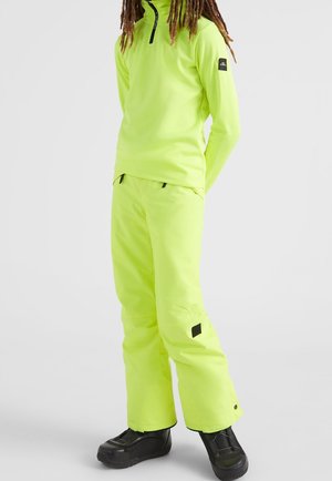 Persona vistiendo una chaqueta y pantalones de esquí de neón amarillo brillante con botas negras, de pie contra un fondo claro y liso.