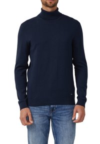 Marineblauer Rollkragenpullover aus weichem Strickstoff, mit langen Ärmeln sowie geripptem Saum und Bündchen. Kombiniert mit blauen Jeans.