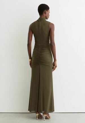 Frau in ärmellosem olivgrünem Maxikleid mit gerafftem Rücken, die an einer weißen Wand steht, trägt beigen Sandalen mit hohen Absätzen und ein goldenes Armband.