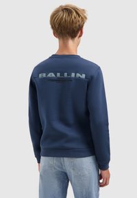 Jongere met kort blond haar, gekleed in een donkerblauwe sweatshirt met de tekst "BALLIN" op de achterkant en lichtblauwe jeans, staand met de rug naar ons gekeerd.