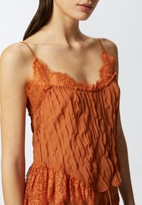 Narančasta teksturirana majica bez rukava s čipkastim obrubom duž dekoltea i rubova, koja ima tanke podesive naramenice i slojeviti volanasti dizajn.