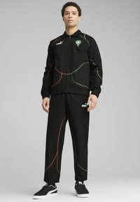 Chándal negro con líneas curvas verdes y rojas, chaqueta con cremallera, puños y cinturilla elásticos. Combinado con zapatillas negras, logo en los pantalones.