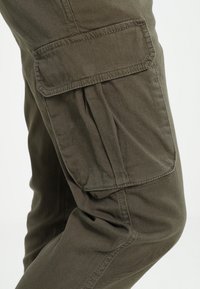 Gros plan sur la jambe d'un pantalon cargo vert olive avec une grande poche à rabat sur la cuisse et des détails de couture visibles.