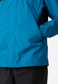 Modrá nepromokavá bunda s hladkou texturou, elastickými manžetami a boční kapsou. Má decentní logo "DRY VENT" poblíž manžety.