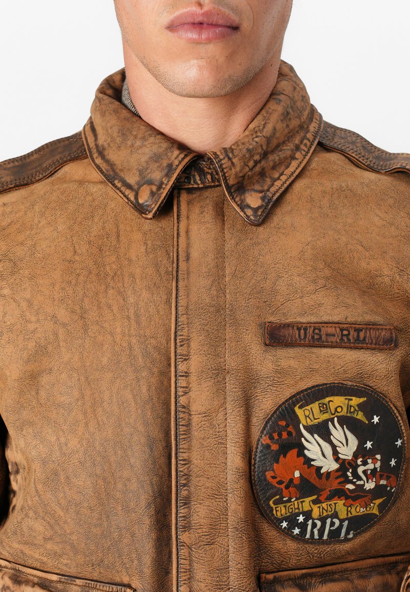 Giacca in pelle marrone invecchiata con un grande patch circolare rappresentante una tigre, ali e "FLIGHT INST." I dettagli includono un colletto con bottoni e una tasca frontale.