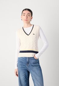 V-Ausschnitt ärmelloser Pullover aus cremefarbenem Kabelstrick mit navyblauen und gelben Streifen am Saum, versehen mit einem kleinen grünen Logo auf der Brust.