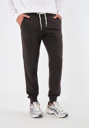 Braune Jogginghosen mit einem Kordelzugbund, vorderen Taschen und schmal zulaufenden Beinen. Getragen mit silbernen Laufschuhen. Glatte Stoffstruktur.