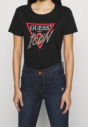 Kvinde iført en sort Guess T-shirt med en rød trekantslogo og "ICON" i rhinsten, kombineret med mørkeblå højtaljede jeans.