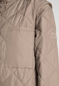 Beige gesteppte Jacke mit Diamantsteppung, großer quadratischer Brusttasche, Knopfverschluss und Stofflogo-Patch am oberen Ärmel.