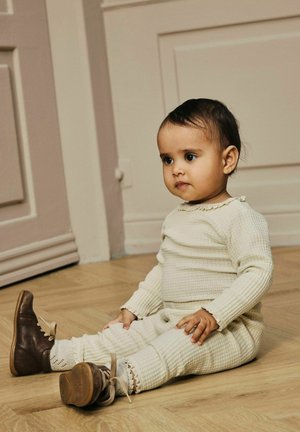 Crème-kleurige gebreide outfit met een gestructureerd patroon, voorzien van gerimpelde randen. Bruine leren schoenen met een strikaccent en lichtgekleurde zolen.