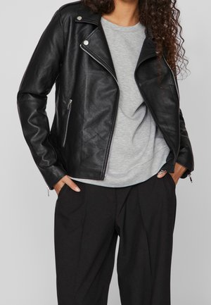 Personne portant une veste de motard en cuir noir par-dessus une chemise gris clair et un pantalon noir, les mains dans les poches.