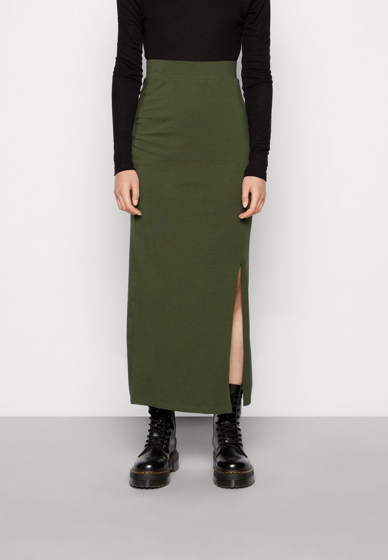 Personne portant une jupe longue taille haute couleur vert olive avec une fente sur le côté, un haut noir à manches longues et des bottes noires à lacets.