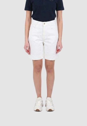 Persona che indossa una maglietta blu navy, pantaloncini bianchi a vita alta fino al ginocchio e scarpe da ginnastica bianche, in piedi davanti a uno sfondo chiaro e uniforme.