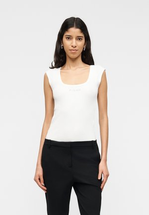 Vrouw met lang donker haar, gekleed in een witte mouwloze top en zwarte broek, staand tegen een effen lichte achtergrond.