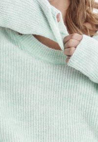 Pull vert clair en maille avec un col ample et des poignets côtelés. Le tissu semble doux et texturé avec une légère transparence.