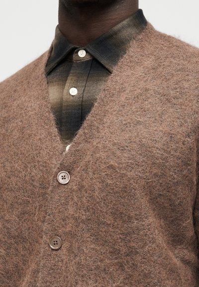 Άνδρας φορώντας καφέ μαλακό πουλόβερ πάνω από ένα πουκάμισο με καρό μοτίβο σε σκουρό καφέ και μπεζ, κοντινή λήψη στο στήθος και στον λαιμό.