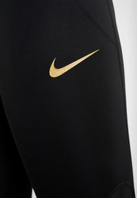 Leggings sportive neri con un design elegante, caratterizzati da un logo Nike dorato glitterato sulla coscia e pannelli in rete testurizzati vicino al polpaccio.