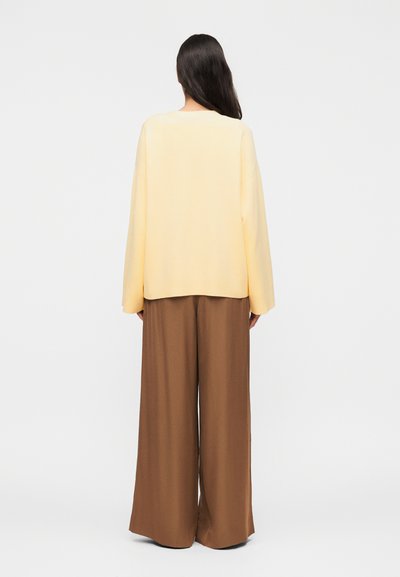 Γυναίκα με μακριά σκούρα μαλλιά, που φοράει ένα άνετο ανοιχτόκίτρινο πουλόβερ και φαρδιά καφέ παντελόνια, στέκεται γυρισμένη πλάτη σε έναν απλό λευκό φόντο.