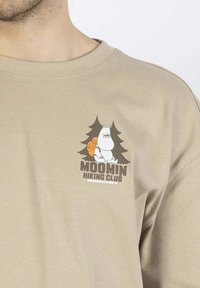 Beige puuvillapaita, jossa on sarjakuvahahmo repun kanssa puiden ympär�öimänä, ja tummanruskea teksti "MOOMIN HIKING CLUB".