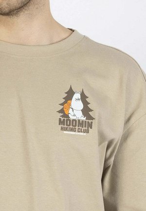 Beige puuvillapaita, jossa on sarjakuvahahmo repun kanssa puiden ympäröimänä, ja tummanruskea teksti "MOOMIN HIKING CLUB".