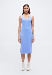 Φωτεινό γαλάζιο βαμβακερό midi φόρεμα με εφαρμοστή γραμμή, λαιμόκοψη σε σχήμα scoop και μπροστινό σήμα με λογότυπο· φορεμένο με λευκά αθλητικά παπούτσια και εκρού κάλτσες αστραγάλου.