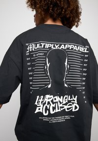 Schwarzes oversized T-Shirt mit einem weißen grafischen Design, das eine Silhouette, Maßstabsmarkierungen und den Text "FALSCH BESCHULDIGT" zeigt.