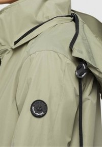 Veste imperméable vert olive à finition mate, avec un patch logo noir sur la manche, des cordons de serrage ajustables et des poches zippées.