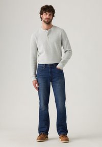 Camisa henley de manga larga gris con tres botones, combinada con jeans rectos azules y zapatos casuales marrones. Diseño simple y funcional.