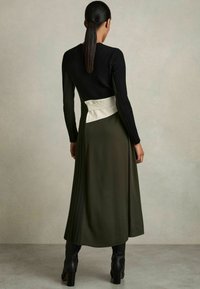Lang jurk met een aansluitend zwart geribbeld lijfje, een soepele olijfgroene rok en een contrasterende crèmekleurige sjerp midden op de taille.