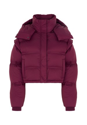 Femme portant une doudoune oversize violet foncé, un pantalon noir à jambes larges, un haut noir, des lunettes de soleil et des chaussures noires, debout devant un fond gris.