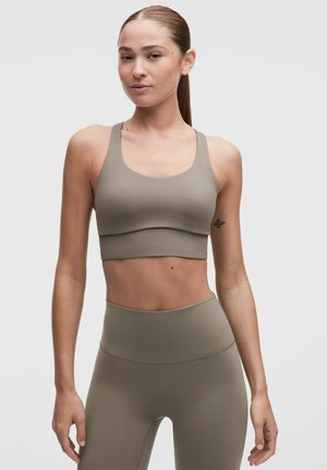Kvinde med langt hår samlet bagpå iført matchende beige sports-bh og høj-taljede leggings, stående mod en ensfarvet baggrund.